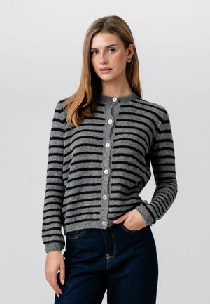 Kvinde iført en grå og sort vandretstribet cardigan med knapper og lange ærmer samt mørkeblå jeans, stående foran en ensfarvet baggrund.