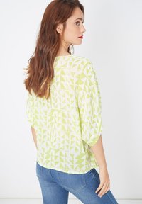 Blusa ligera y transparente con un patrón geométrico en un amarillo-verde brillante sobre un fondo blanco. Corte holgado con mangas amplias.