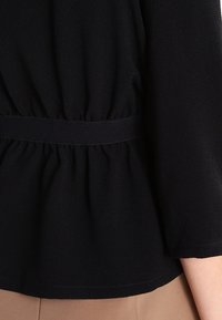Zwarte blouse met een geplooide textuur bij de taille, voorzien van een gladde stof en een brede, zachte tailleband. Lange, licht uitlopende mouwen zijn zichtbaar.