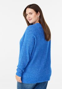 Kobieta z długimi brązowymi włosami, nosząca srebrne kolczyki, jasnoniebieski sweter i niebieskie dżinsy, spoglądająca przez ramię, uśmiechająca się.