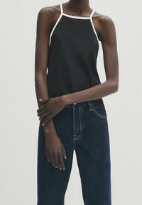 Personne mince portant un haut sans manches noir avec bord blanc et un jean taille haute bleu foncé, debout avec les bras croisés.