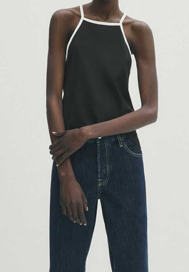 Personne mince portant un haut sans manches noir avec bord blanc et un jean taille haute bleu foncé, debout avec les bras croisés.