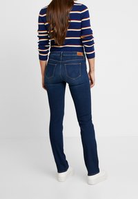 Jeans de mezclilla azul oscuro con un diseño ajustado, que cuentan con dos bolsillos traseros y una etiqueta de cuero en la cinturilla. Se combinan con zapatos blancos.