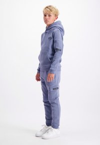 Blonde jongen die staat, in een blauwe hoodie en bijpassende sweatpants met zijzakken, en witte sneakers, op een witte achtergrond.