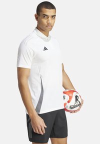 Weißes Polohemd mit Kragen, verziert mit grauen Akzenten und dem Adidas-Logo. Schwarze Shorts und ein rot-weißes Fußball, der in der Hand gehalten wird.