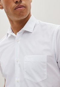Cămașă albă cu mâneci scurte, confecționată dintr-un material neted, având un guler ascuțit și o buzunare pe partea stângă a pieptului. Design clasic cu nasturi.
