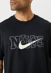 Černé bavlněné tričko s velkým, stylizovaným logem "NIKE" v šedé a bílé, s výrazným grafickým prvkem swoosh. Design s kulatým výstřihem.
