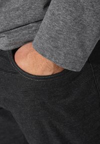 Graues Langarmhemd, das über schwarzen Textilhosen mit sichtbarem Tascheneingriff, verstärkten Nähten und subtilen horizontalen Mustern getragen wird.