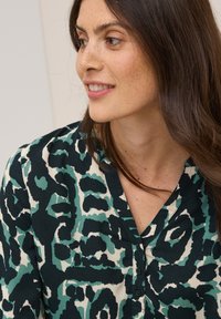 Blouse avec un imprimé léopard abstrait vert et noir, décolleté en V et détails de boutons. Fabriquée en tissu léger avec une texture douce.