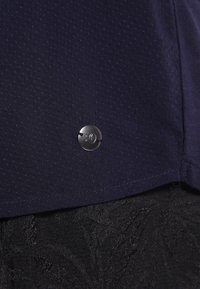 Tissu texturé bleu foncé avec un petit logo circulaire en métal cousu près de l'ourlet inférieur sur un tissu noir à motifs en dessous.