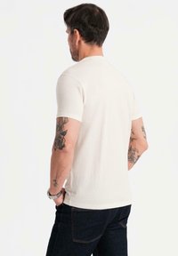 Kremowa bawełniana koszulka z krótkimi rękawami i okrągłym dekoltem, charakteryzująca się luźnym krojem i gładką fakturą. Noszona z ciemnoniebieskimi dżinsami.