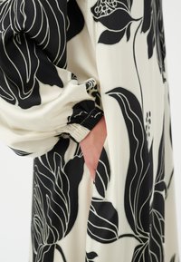 Robe fleurie à base crème avec de grands motifs floraux noirs. Caractérisée par des manches longues froncées et un détail de poche sur le côté. Texture lisse.