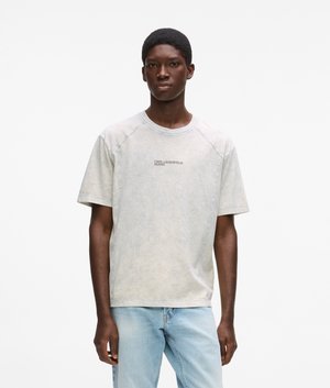 Mlad moški stoji ob belem ozadju, oblečen v svetlo oprano majico s kratkimi rokavi in svetlo modre kavbojke.