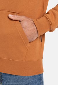 Orange hættetrøje med frontlomme, ribbede manchetter og nederkant. Stoffet ser blødt ud med en subtil tekstur. Synlig logomærkat på siden.