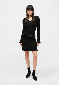Black ribbed sweater dress with long sleeves and flared cuffs, cinched at the waist with a black belt. Styled with black socks and pointed shoes.