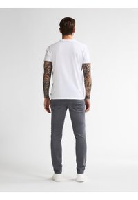 Camiseta blanca de manga corta, jeans ajustados grises y zapatillas blancas. Tatuajes visibles en los brazos. Diseño simple sin patrones ni logotipos visibles.