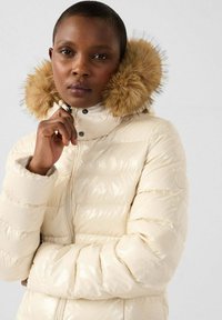 Giacca puffer crema con texture lucida e cappuccio con pelliccia. Presenta una chiusura con zip e un design trapuntato. Morbida, calda e alla moda.