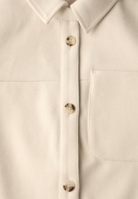 Jasnobeżowa koszula zapinana na guziki, wykonana z miękkiego materiału, z kołnierzykiem w kształcie szpica, sześcioma guzikami i kieszenią na lewej piersi.
