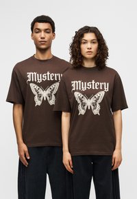 Καφέ βαμβακερά μπλουζάκια με μεγάλα λευκά γραφικά πεταλούδας και τη λέξη «Mystery» σε διακοσμητική γραμματοσειρά. Κοντά μανίκια, άνετη εφαρμογή.
