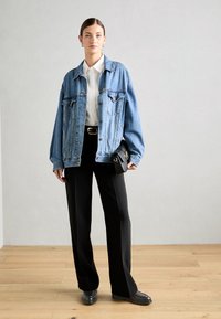 Chaqueta de mezclilla en azul claro con corte oversize, botón delantero y dos bolsillos en el pecho, combinada con pantalones de pierna ancha negros y mocasines negros.