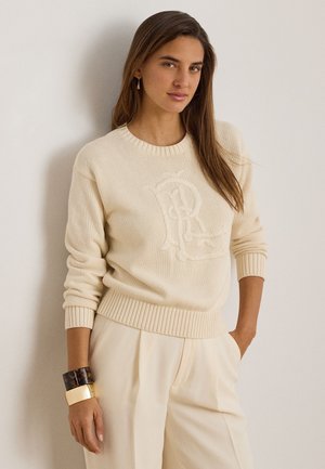 Kvinde med langt brunt hår iført cremefarvet strikket sweater med præget logo, cremefarvede bukser, guld- og skildpaddeskalsarmbånd, stående mod en væg.