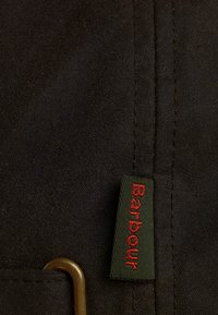 Țesătură neagră cu cusături vizibile, având un etichetă verde cu text roșu "Barbour". De asemenea, este prezent un element de fixare de culoare bronz.