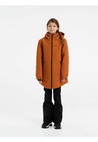 Prenda exterior acolchada naranja con capucha, cierre frontal de cremallera y dos bolsillos laterales, llevada sobre pantalones de esquí negros y botas.
