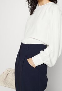 Mujer vistiendo una blusa blanca de manga larga metida dentro de unos pantalones de tiro alto azul marino, con una mano en el bolsillo, sosteniendo un bolso de mano tejido beige.