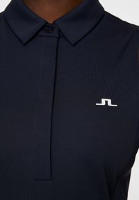 Marineblå polo skjorte med krave, der har en frontplacket og et lille hvidt logo på venstre bryst. Fremstillet af glat, strækbart stof.