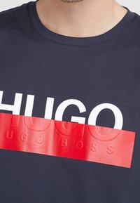T-paita laivastonsinistä crew neck -mallia, jossa on suuri valkoinen "HUGO" -logo kiiltävän punaisen nauhan päällä. Logossa on teksturoituja accentteja brändin nimestä.