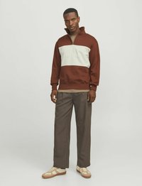 Barna és krémszínű cipzáros sweatshirt színblokkokkal, textúrázott betűzéssel. Szürke bő nadrággal és bézs sportcipővel párosítva.