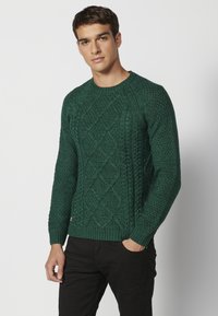 Groene gebreide trui met een ruitenpatroon, ronde halslijn en geribbelde mouwen en zoom. G gedragen met zwarte broek.