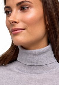 Maglione grigio a collo alto con texture a costine, caratterizzato da un colletto alto e una finitura liscia, incorniciato da capelli sciolti e trucco naturale.