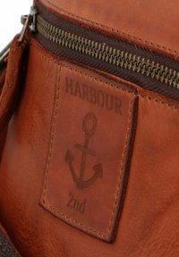 Bruine leren tas met een rits en een gegraveerd ankerlogo etiket, met een gladde textuur en zichtbare stiksels.