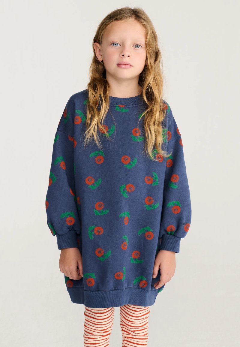 Jeune fille aux longs cheveux blonds, portant un sweat-shirt bleu oversize avec des imprimés de fleurs orange et des leggings rayés rouges et blancs.