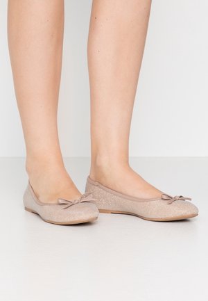 Stopy w błyszczących, beżowych baletkach z małymi kokardkami, stojące na gładkiej białej powierzchni na tle białego tła.