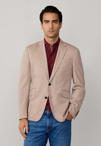 Hellbeige Blazer mit zwei Vorder pockets, Ein-Knopfverschluss und Reverskragen, kombiniert mit einem burgunderfarbenen Poloshirt und blauen Jeans.