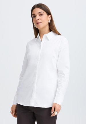 Femme aux cheveux bruns et raides, portant des boucles d'oreilles anneaux argentées, une chemise blanche boutonnée et un pantalon noir, debout devant un fond clair.