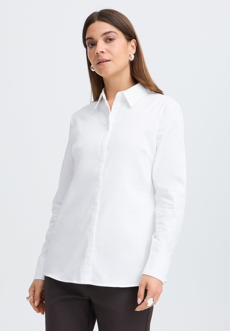 Femme aux cheveux bruns et raides, portant des boucles d'oreilles anneaux argentées, une chemise blanche boutonnée et un pantalon noir, debout devant un fond clair.