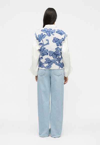 Femme aux longs cheveux foncés portant une chemise blanche à manches longues, un gilet tricoté bleu à fleurs et un jean bleu clair à jambes larges, debout de dos sur un fond uni.