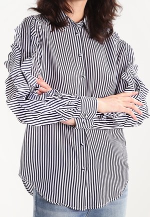 Kobieta ubrana w czarno-białą, pionowo prążkowaną, długorękawową koszulę z marszczeniami na rękawach, z założonymi na siebie ramionami, w połączeniu z niebieskimi dżinsami.