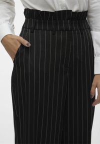 Svarta randiga byxor med en volangmidja, sidofickor och en mjuk yta; smala vita ränder skapar ett kontrasterande mönster.