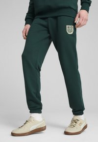 Tamsiai žali sportiniai kelnių, prigludusio silueto, su kreminės spalvos logotipo aplikacija šlaunyje ir elastingais rankogaliais. Suderinti su smėlio spalvos sportbačiais.