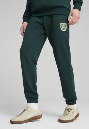 Pantalones de chándal verde oscuro con un diseño ajustado, que cuentan con un logo crema en el muslo y puños elásticos. Combinados con zapatillas beige.