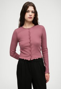 Cardigan à manches longues côtelé en rose poussiéreux, avec un col arrondi, des boutons sur le devant et un ourlet festonné, associé à un pantalon noir.