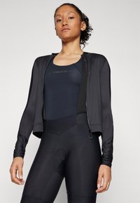Schwarze Radjacke mit langen Ärmeln und einem Reißverschluss vorne, getragen über einem dunklen Bodysuit, mit glatter Textur und atmungsaktivem Material.