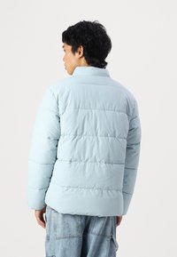 Light blue puffer jacket with a high collar, featuring quilted lines and a relaxed fit. Worn over loosely fitted blue denim pants.