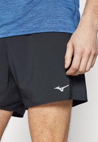 Schwarze Sportshorts mit glatter Textur haben ein weißes Logo am Saum. Ein blaues, strukturiertes Hemd ist oberhalb der Shorts sichtbar.