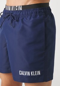 Shorts de baie bleumarin cu cordon de ajustare, având o bandă neagră cu inscripția "CALVIN KLEIN" și logo-ul alb al brandului pe tiv. Textură netedă.
