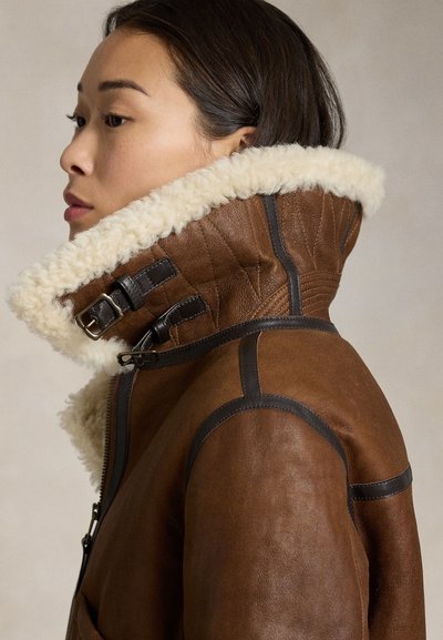 Woman in brown leather jacket with high shearling collar and black buckle straps, shown in side profile against plain background.